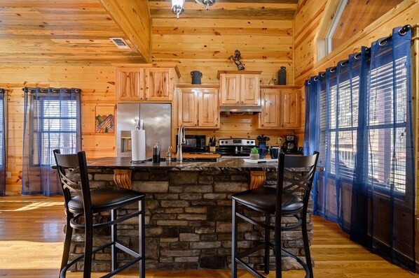 A rustic kitchen with wooden cabinets, a stone island, and a bar area with stools.