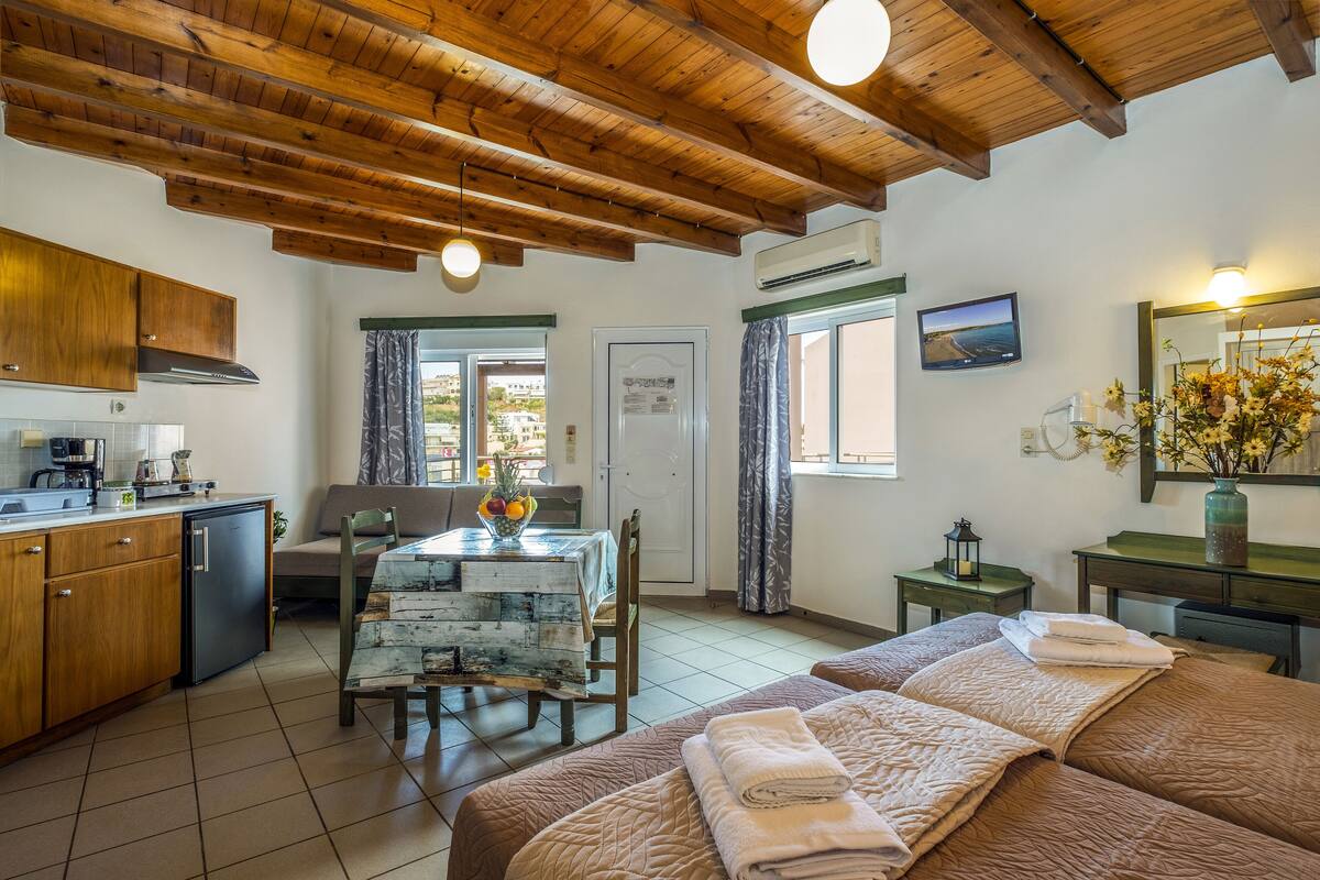 A room with wooden beams, a kitchen with wooden cabinets, a dining table with a fruit bowl, a bed with towels, and a TV on the wall.