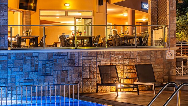 A pool area with a stone wall, lounge chairs, and a restaurant with outdoor seating. - Theos village apartments (Chania)