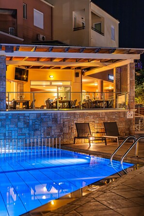 A pool area with a stone wall, lounge chairs, and a restaurant with outdoor seating. - Theos village apartments (Chania)