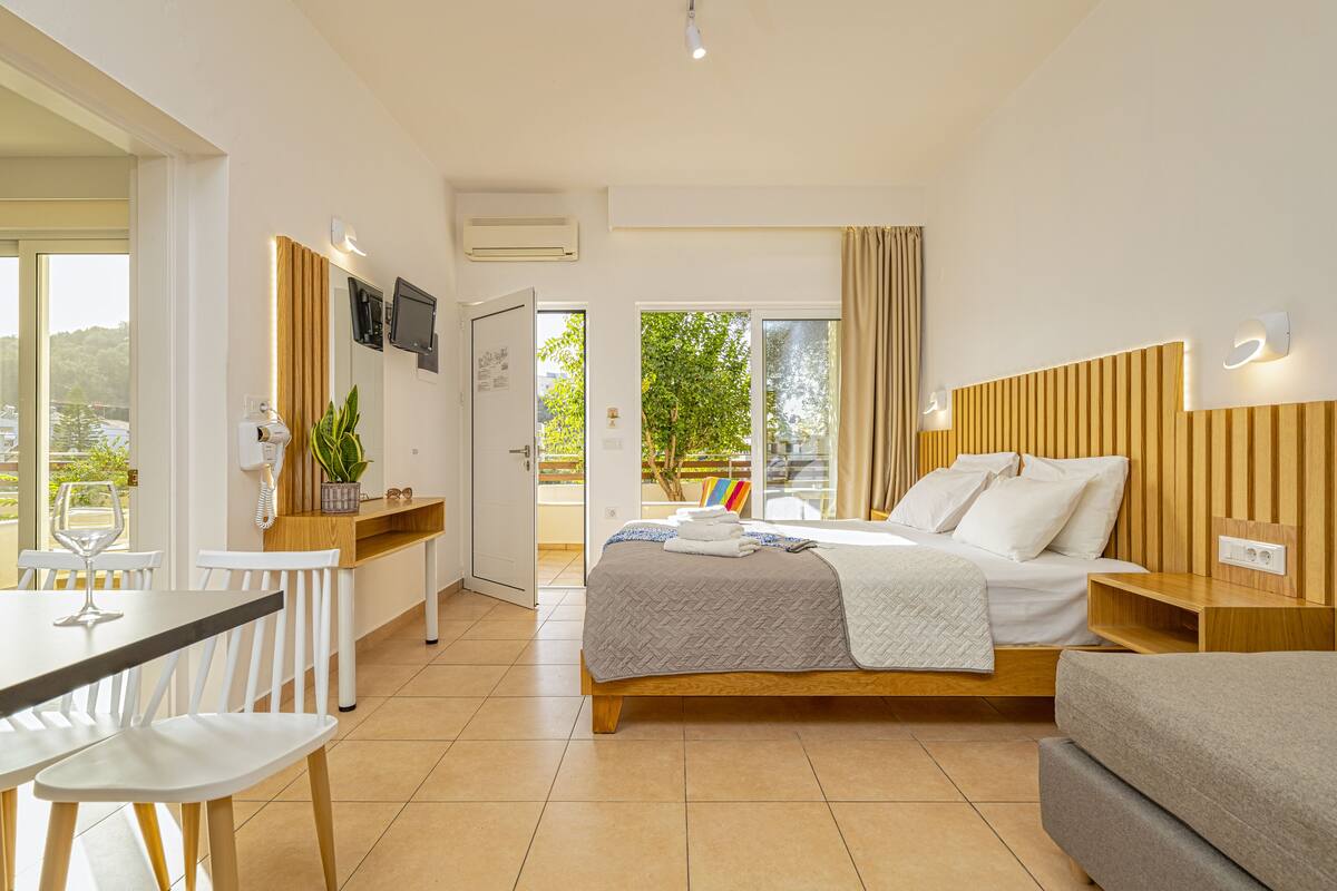 A modern hotel room with a bed, a desk, a dining table, and a balcony with a view.