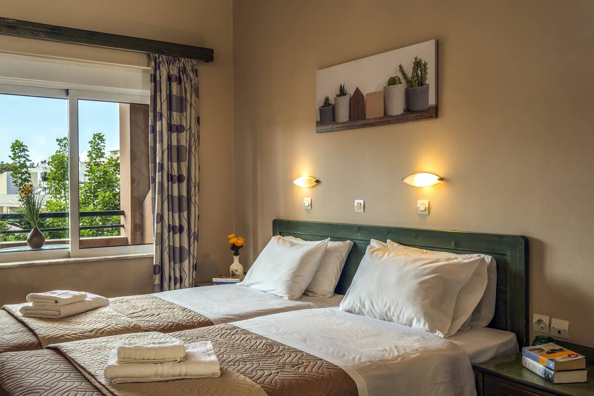 A hotel room with two beds, a window with curtains, a nightstand with a lamp, and a wall-mounted shelf with potted plants.