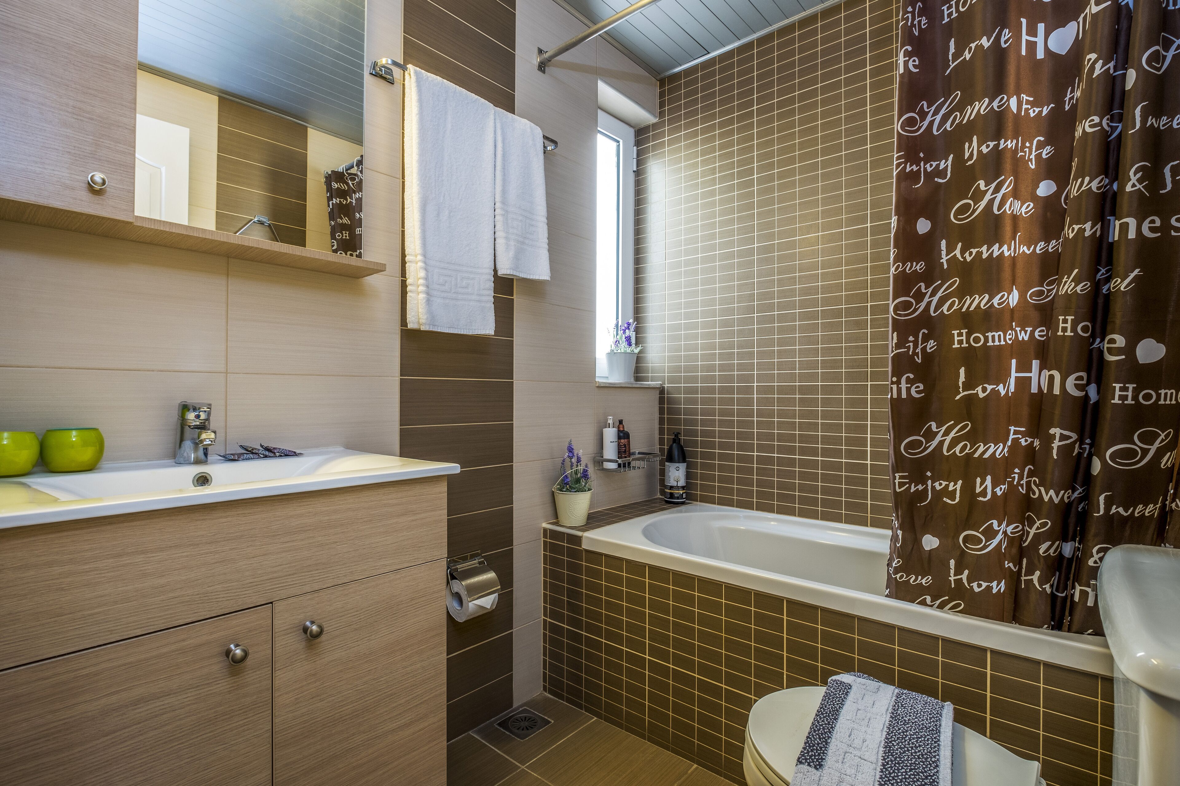 Ein modernes Badezimmer mit einer Badewanne, einer Toilette und einem Waschbecken. Über dem Waschbecken befindet sich ein Handtuchhalter und ein Fenster mit Jalousien.