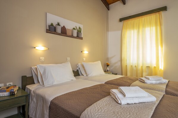 A hotel room with two beds, a nightstand with a book, a wall-mounted shelf with plants, and a window with curtains. - Theos village apartments (Chania)