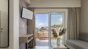 A modern living room with a sofa, a coffee table, and a TV on the wall. There is a balcony with a view of the city and a door leading to another room. - Theos village apartments (Chania)