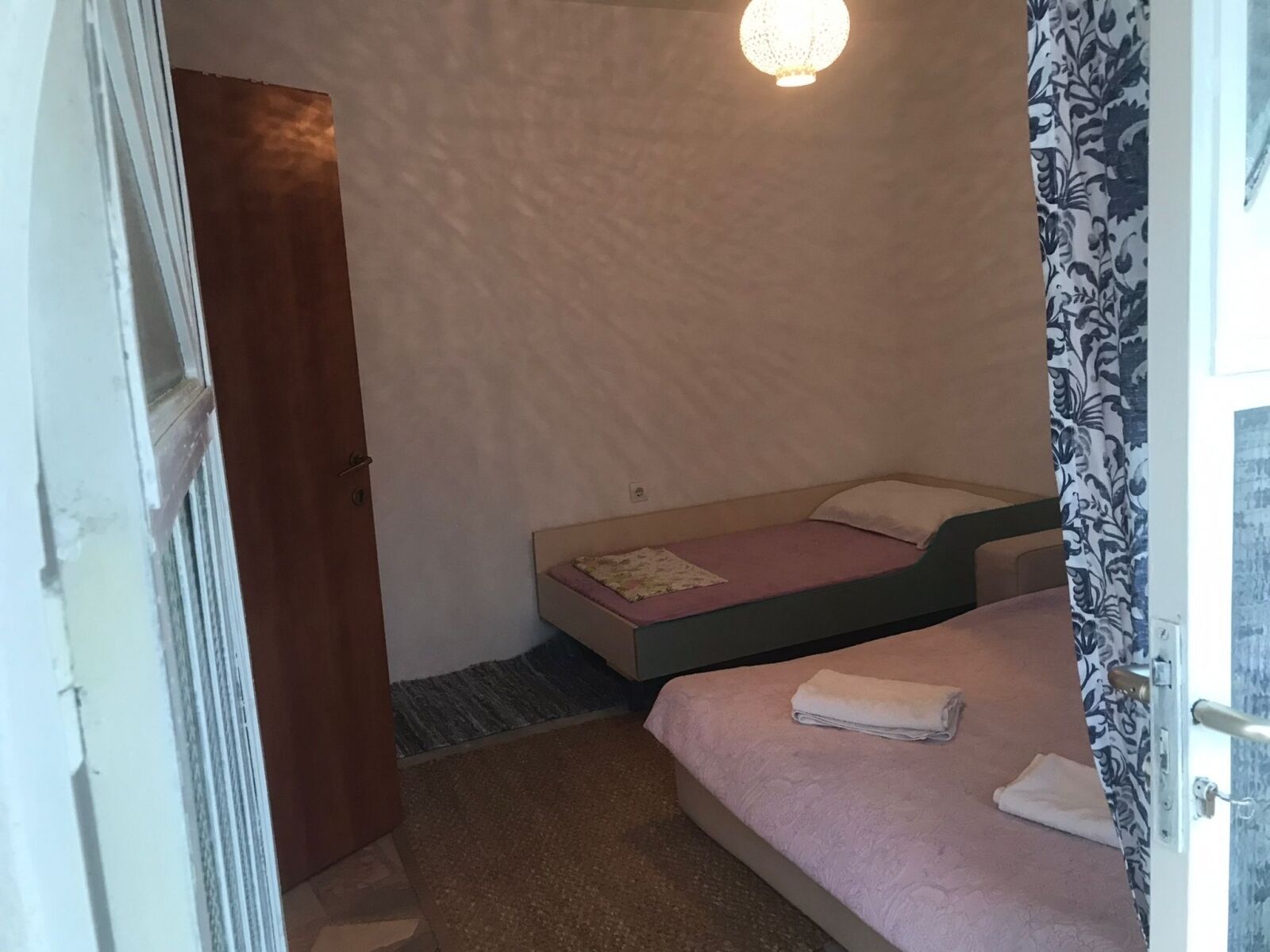 Una piccola camera con un letto singolo, una porta in legno, una tenda decorata e una luce a soffitto.
