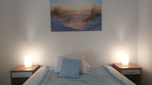 Et soveværelse med en seng, to sengeborde med lamper, og et maleri af en strand på væggen.
