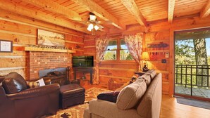 A cozy log cabin interior with a fireplace, leather sofas, a television, and a large window with curtains. - Stunning Mountain Views!!! Secluded Log Cabin with Hot Tub near West Jefferson  (Lansing)