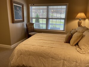 Une chambre avec un grand lit, une chaise, une fenêtre avec des stores et une lampe.