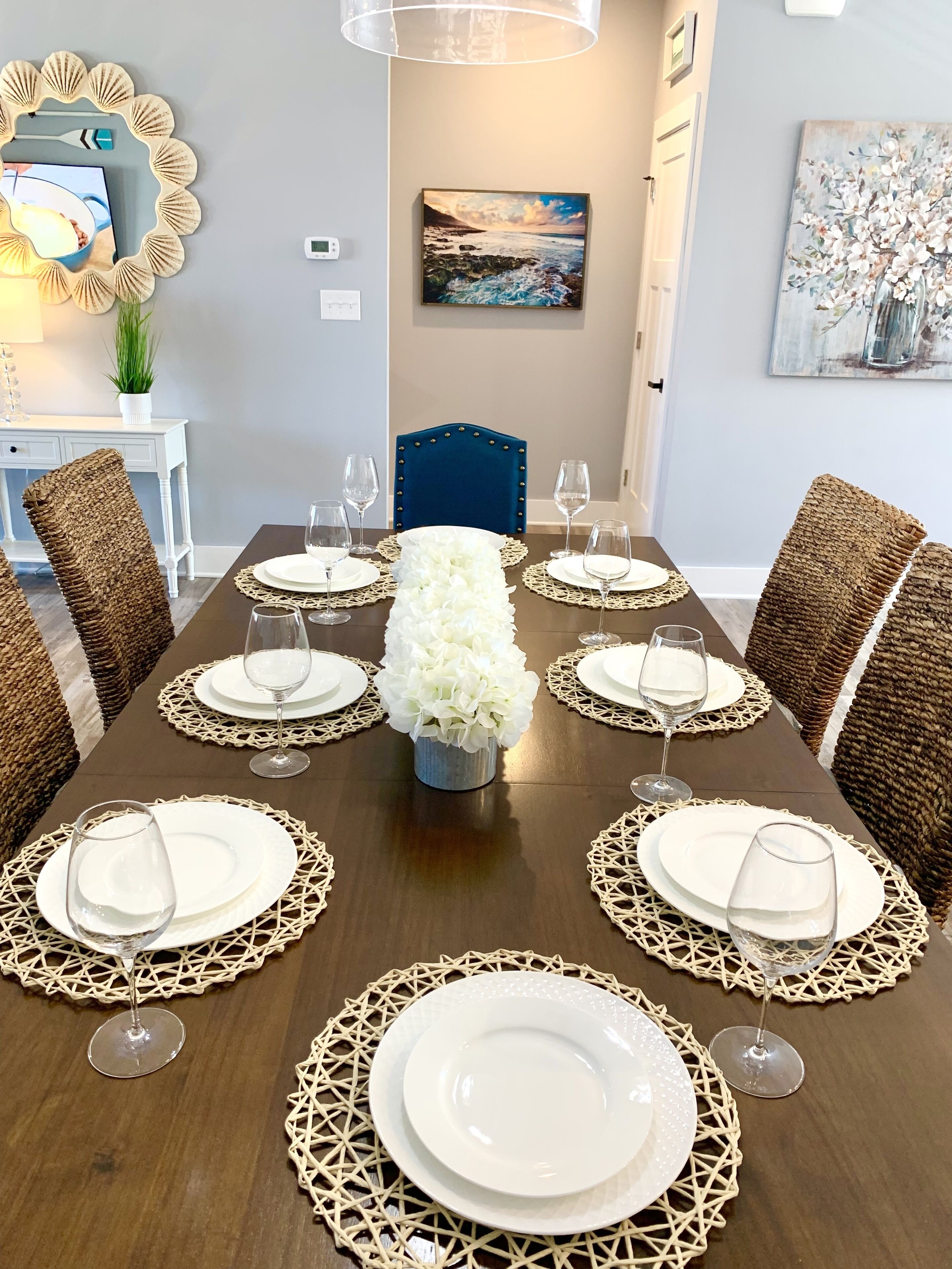 A dining table set with white plates and wine glasses, surrounded by wicker chairs, with a vase of white flowers in the center.