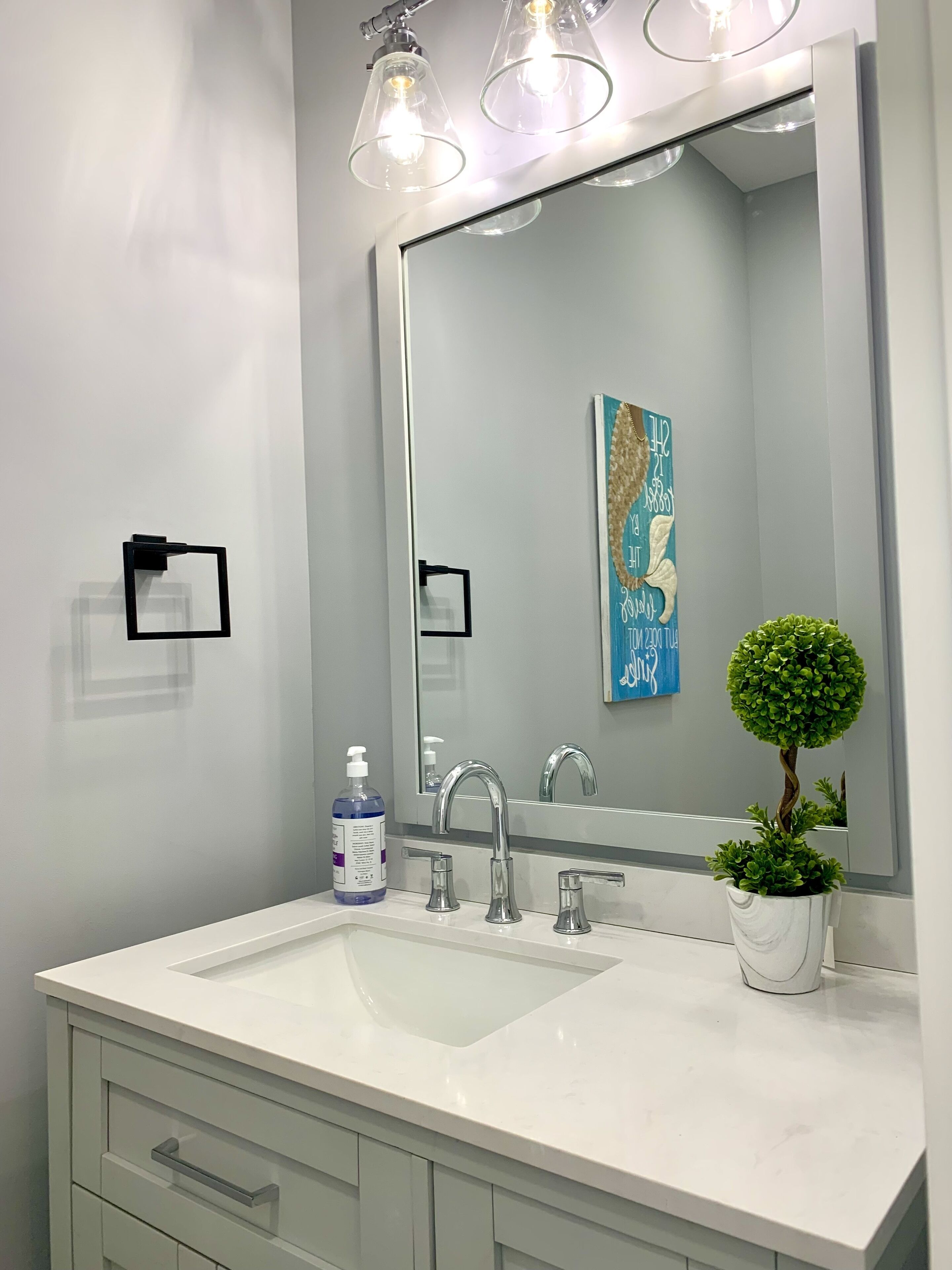 A modern bathroom with a white sink, two faucets, a large mirror, and a small potted plant.