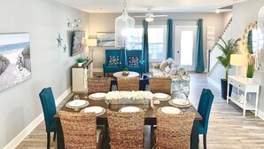 A dining area with a large table set for six, surrounded by wicker chairs. There is a blue sofa and a white console table with a lamp in the background. - IslandCoveParadise: Luxury-NewlyBuiltTH! OnTheWater-Boardwalk-Pool-w/2 CarGarage (Ocean City)