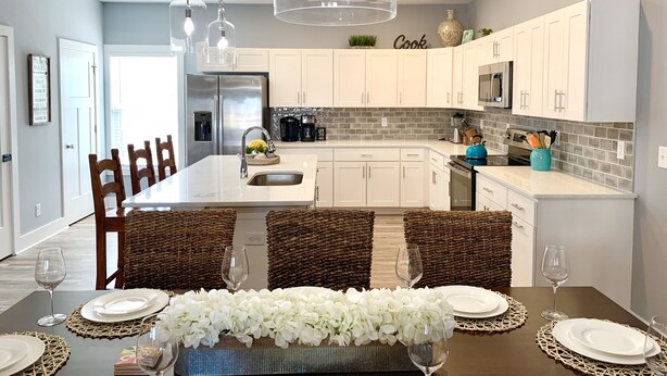A modern kitchen with white cabinets, a stainless steel refrigerator, a built-in oven, and a microwave. There is a dining area with a table set for two, featuring wicker chairs and a centerpiece of white flowers. - IslandCoveParadise: Luxury-NewlyBuiltTH! OnTheWater-Boardwalk-Pool-w/2 CarGarage (Ocean City)
