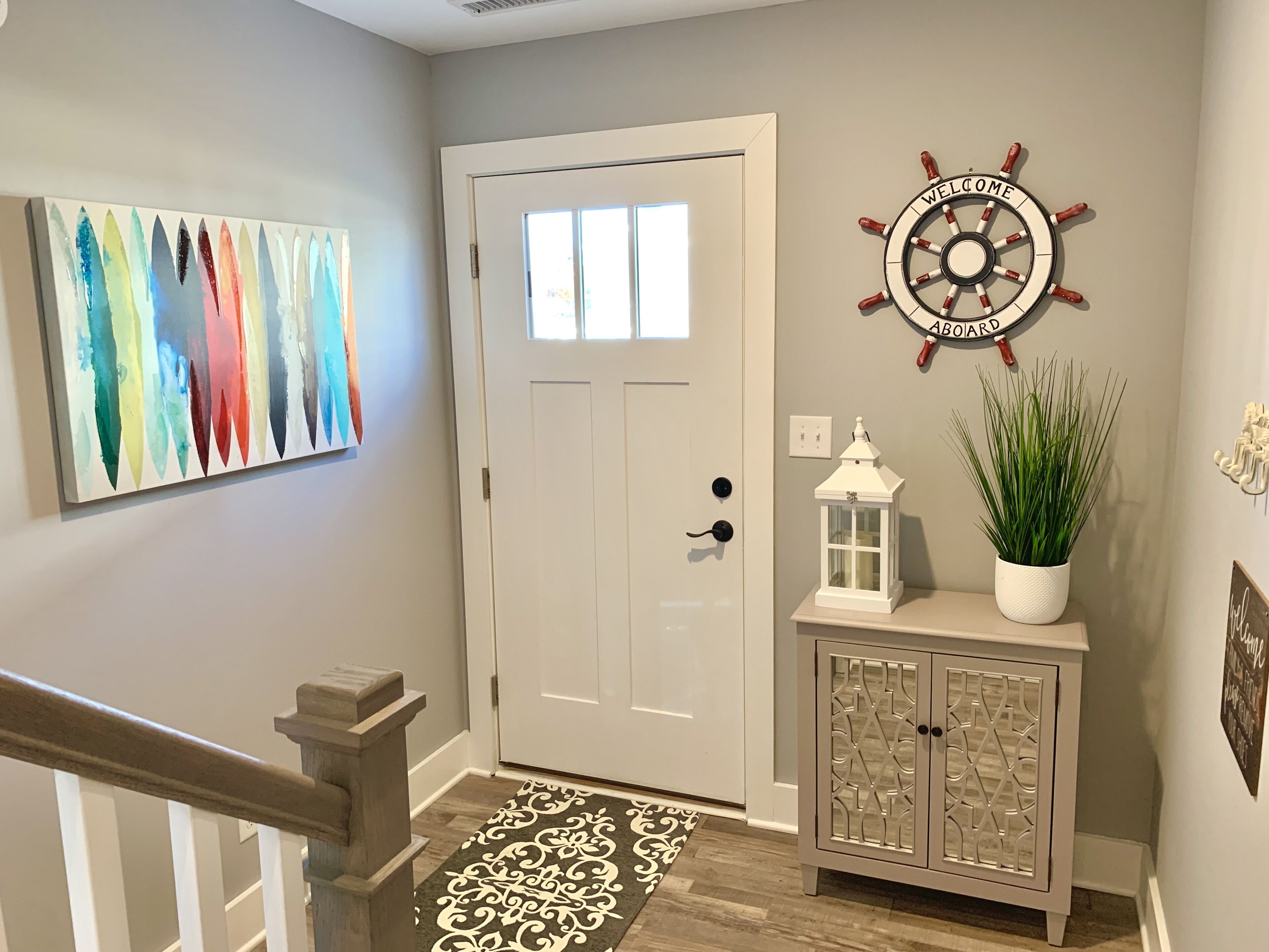 Un couloir avec une porte blanche, un panneau décoratif représentant un gouvernail de bateau fixé au mur, une peinture abstraite aux couleurs vives, une plante en pot et un meuble décoratif.