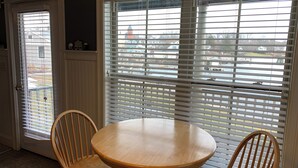 Un coin repas avec une table ronde et quatre chaises en bois, de grandes fenêtres équipées de stores et une vue sur un quartier résidentiel.