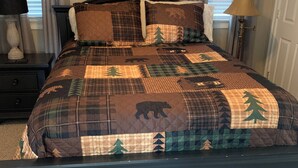 Un lit recouvert d’une courtepointe ornée de motifs représentant une forêt et des animaux, une table de chevet avec une lampe, et un tableau encadré au-dessus du lit.