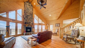 A spacious living room with a stone fireplace, wooden ceiling, and large windows offering a view of the outdoors. - A True Montana Cabin with Sweeping Views of the Mountains and the Madison River. (Cameron)