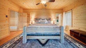 A wooden cabin bedroom with a bed, two bedside lamps, a ceiling fan, and a window with blinds. - A True Montana Cabin with Sweeping Views of the Mountains and the Madison River. (Cameron)
