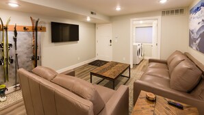 Un salon avec deux canapés en cuir, une table basse, un téléviseur fixé au mur, des skis et du matériel de ski appuyés contre le mur, et un tableau représentant une montagne accroché au mur.