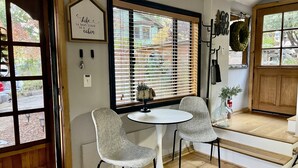 A cozy room with a wooden door, a large window with blinds, two grey chairs around a small white table, and a small potted plant on the windowsill.