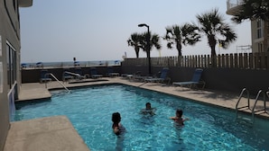 A swimming pool with lounge chairs and palm trees, surrounded by a fence and buildings.