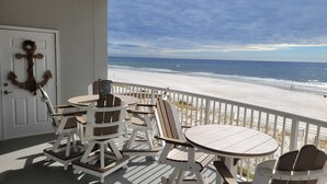 A beachside balcony with white furniture, including a round table and two chairs, overlooking a sandy beach and the ocean.