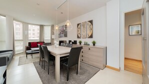 A modern dining area with a large table, chairs, and pendant lighting. There are clocks on the wall and a sofa in the background. - Apartment (Madrid)