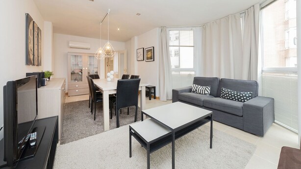 A modern living room with a grey sofa, a dining table with chairs, a TV, and a large window with curtains. - Apartment (Madrid)