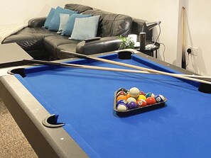 A pool table with a set of billiard balls in the center, a pool cue, and a black leather couch with blue cushions in the background.
