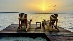Une terrasse en bois avec deux fauteuils Adirondack donnant sur un coucher de soleil au-dessus d’une étendue d’eau.