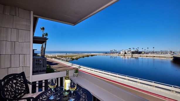 A balcony with a view of the beach and marina, featuring a table with wine glasses and a bottle. - Panoramic Ocean, Marina & River Views - Steps to Beach & Harbor (Oceanside)