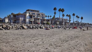 A beach with a rocky shoreline, palm trees, and a row of multi-story buildings with balconies. - Panoramic Ocean, Marina & River Views - Steps to Beach & Harbor (Oceanside)