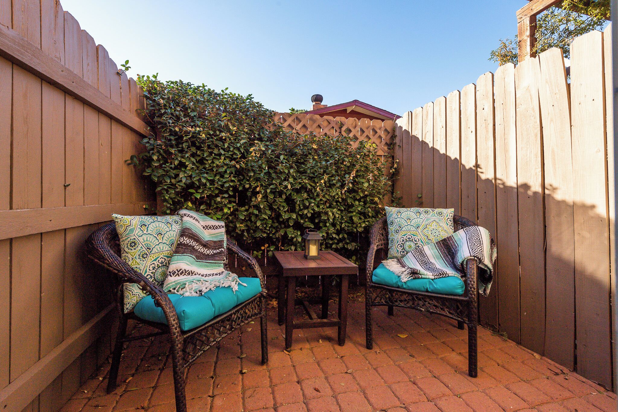 Une terrasse avec deux fauteuils en osier dotés de coussins à motifs, une petite table sur laquelle est posée une lanterne, et une clôture en bois.