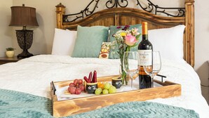 Una cama bien hecha con cabecera de madera, una bandeja con frutas y una botella de vino, y una mesita de noche con una lámpara.