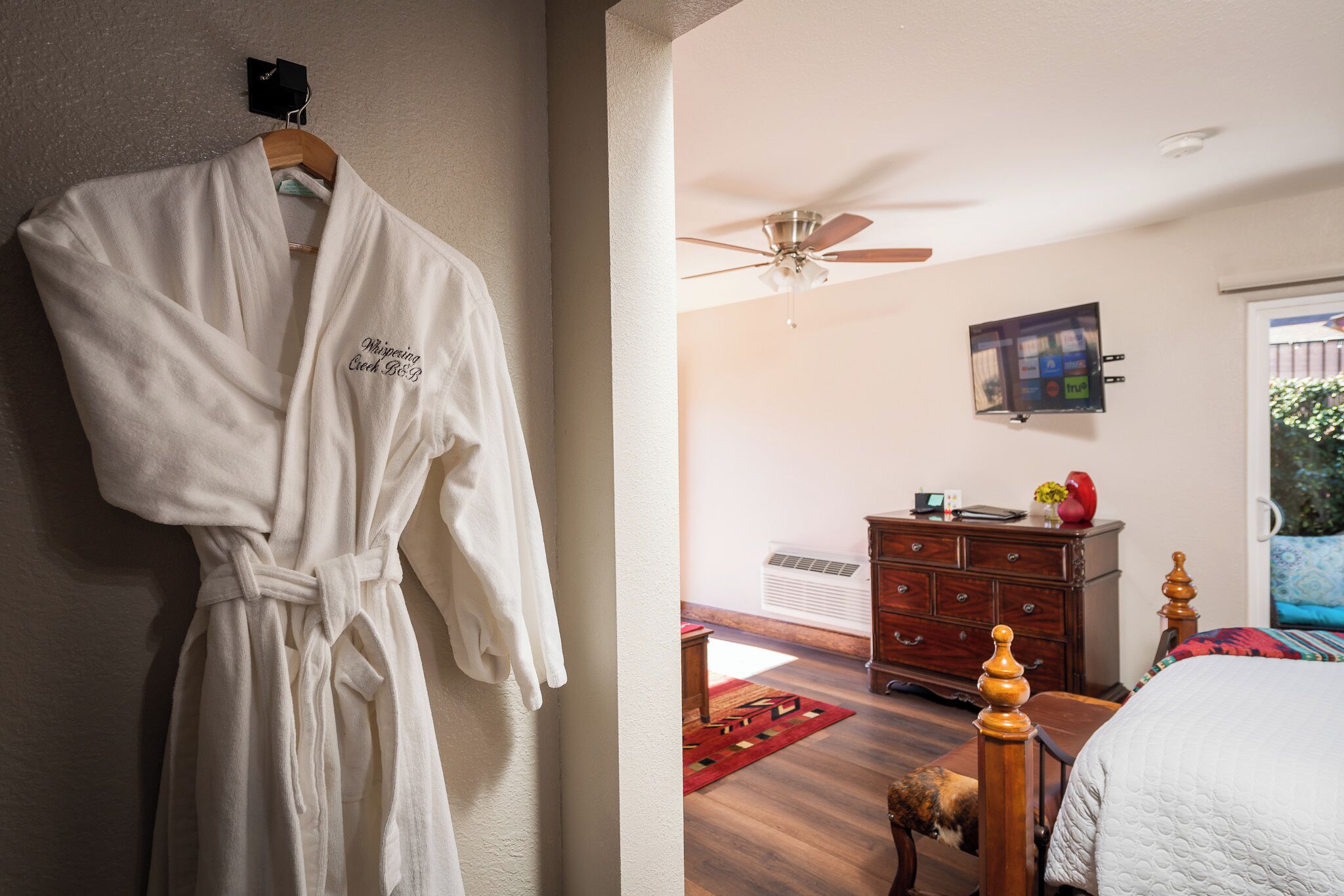 Una bata blanca colgada en la pared, un tocador de madera, una cama con colcha blanca, un ventilador de techo y un televisor montado en la pared.