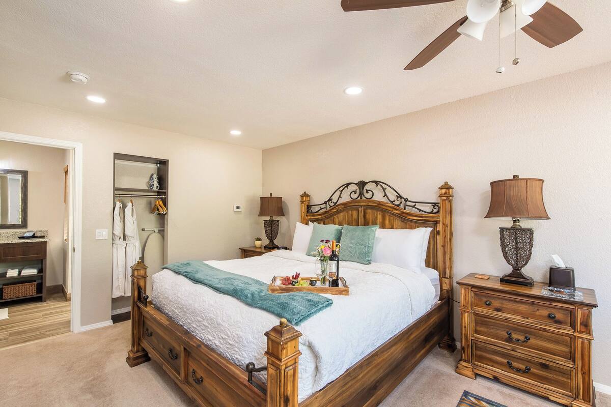 Un dormitorio ordenado con una cama de madera, mesitas de noche, un ventilador de techo y una mesita auxiliar con lámpara.