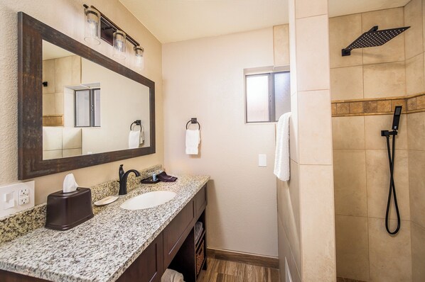 A bathroom with a granite countertop, a large mirror, a towel rack, a shower with a handheld showerhead, and a window. - Whispering Creek B&B (Sedona)