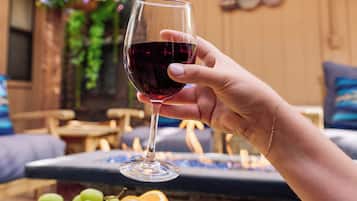 A wooden table with a glass of red wine, a wooden board with fruits and cheese, and a cozy outdoor seating area with cushions and a hanging plant.