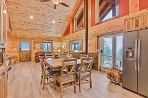 A wooden cabin interior with a dining area, kitchen, and large windows.