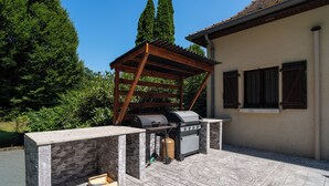 An outdoor barbecue area with a stone and wood structure, a grill, and a gas tank. - Charming country house in a wooded garden in Perigord Vert (La Coquille)