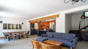 A modern living room with a sectional sofa, dining table, and chairs, a kitchen in the background, and a staircase leading to an upper level. - Charming country house in a wooded garden in Perigord Vert (La Coquille)