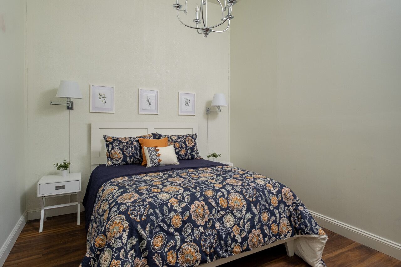 Un dormitorio con cama, mesitas de noche, lámparas de pared y cuadros enmarcados.