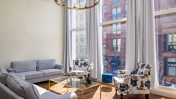 A modern living room with a grey sofa, a floral armchair, a small round table, and a large window with a view of red brick buildings. - Gold Suite by Hollyhock. Executive-Level Living (Louisville)