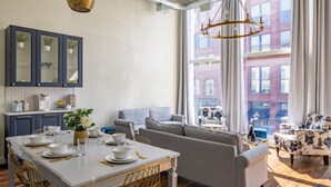 A modern kitchen with a dining area, a sofa, and a view of a red brick building. - Gold Suite by Hollyhock. Executive-Level Living (Louisville)
