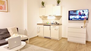 En moderne stue med en sofa, et sofabord med en bærbar computer, et køkkenområde med skabe og et fjernsyn der viser et program, samt en lille plante på disken.