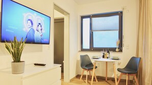 En moderne stue med et fladskærms-tv, en stueplante, et spisebord med stole og et vindue med persienner.