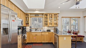 A modern kitchen with wooden cabinets, a stainless steel refrigerator, a dining area with a glass table, and a view of the outside through large windows.
