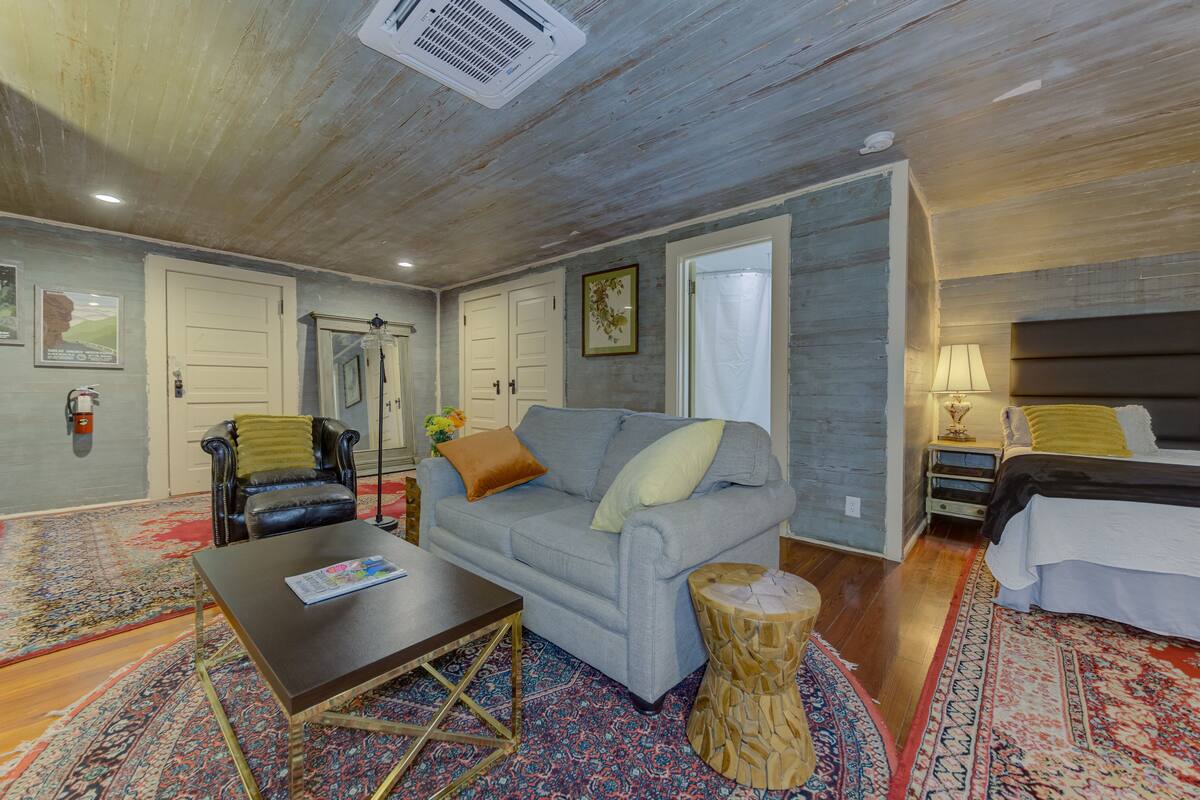 A room with a grey sofa, a wooden coffee table, a patterned rug, a small wooden stool, and a bed with a white duvet.