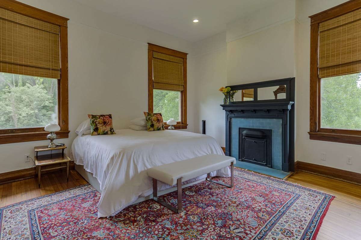 A bedroom with a large bed, a bench, a fireplace, and a window with a view of trees.