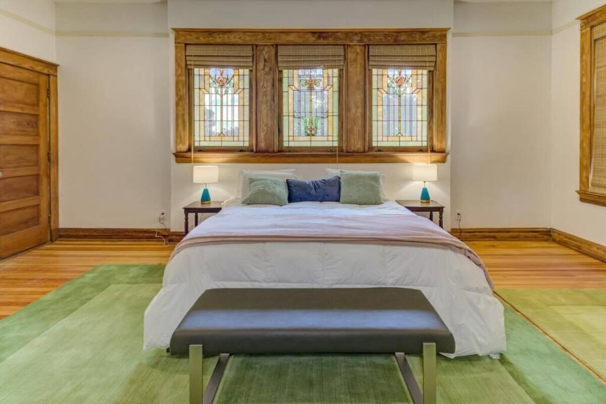 A bedroom with a bed, two nightstands, a bench, and a window with stained glass.